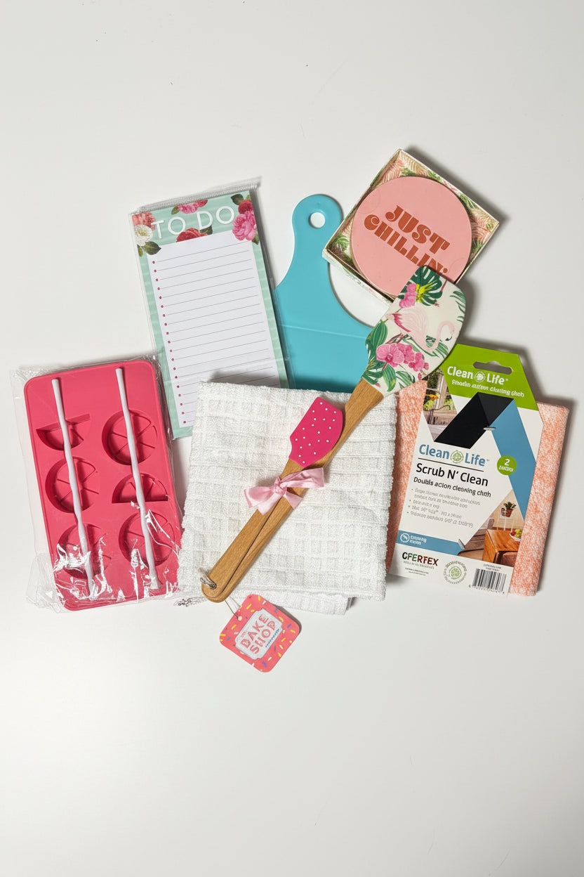 A flat-lay collection of various small household and kitchen accessories on a dark green textured background, including a pink silicone mold, a white towel, a wooden spatula, a notepad, a blue cutting board, and a package of Clean W Clean scrub.