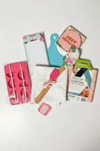 A flat-lay collection of various small household and kitchen accessories on a dark green textured background, including a pink silicone mold, a white towel, a wooden spatula, a notepad, a blue cutting board, and a package of Clean W Clean scrub.